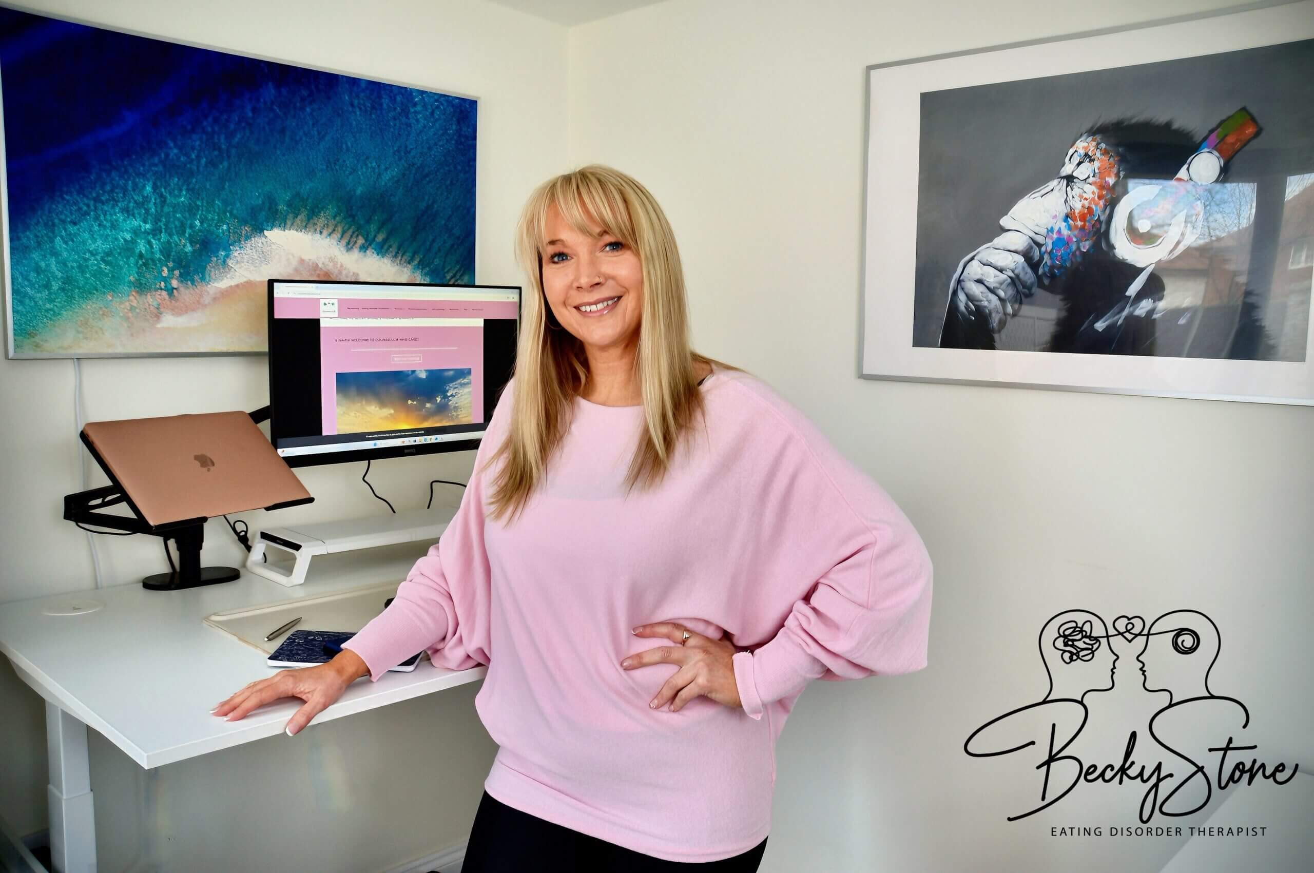 Becky Stone | Eating Disorder Therapist | Online Support & Recovery Guidance Becky Stone, Eating Disorder Therapist, smiling in her modern office with a dual-screen setup displaying her website, offering online support and therapy for eating disorders.